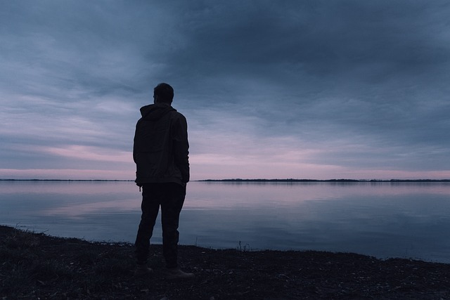 Eine Person steht ganz alleine am Wasser und schaut in den dunkelwerdenden Himmel übers Wasser.