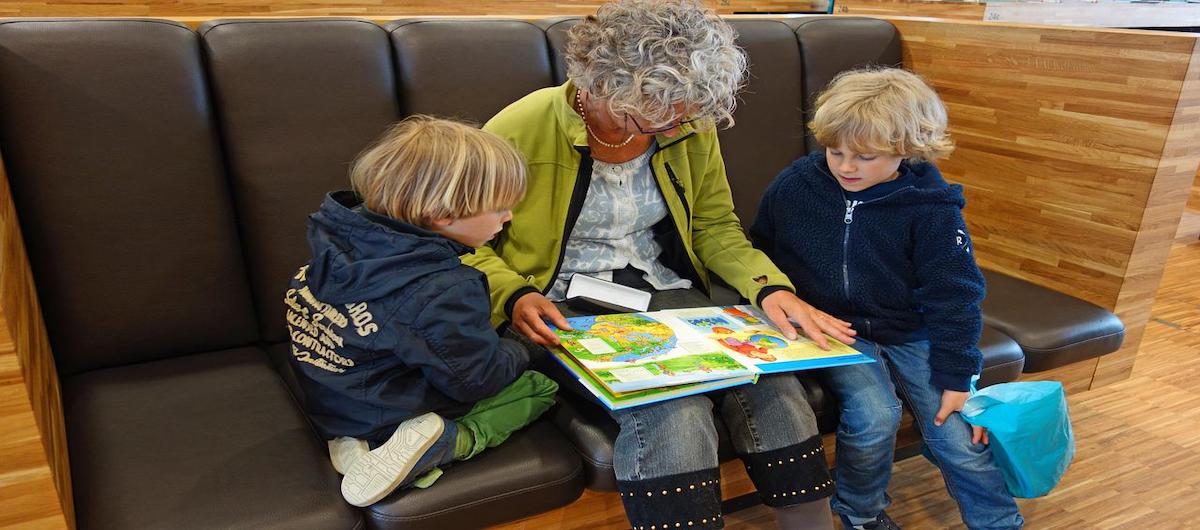 Eine Frau sitzt auf einem braunen Sofa, zwei Kinder links und rechts neben ihr. Die Frau liest aus einem Buch den Kindern vor.